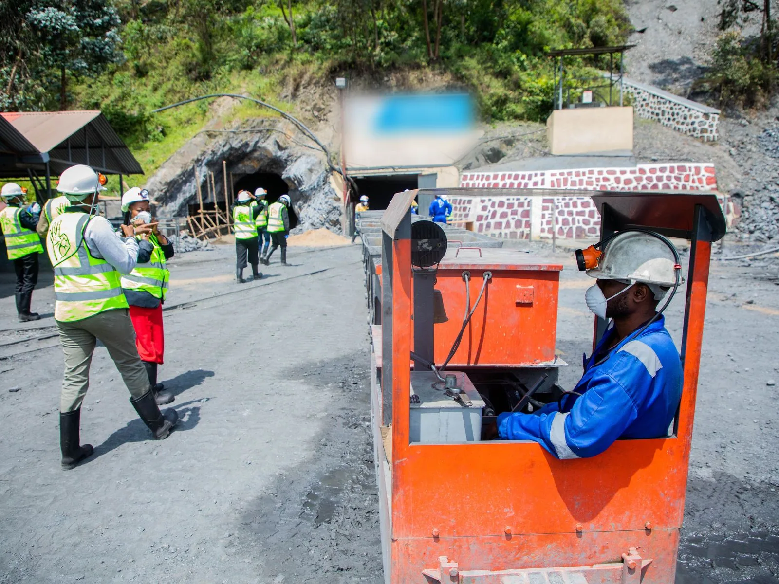 Miners in hard hats and high-visibility vests at a tunnel entrance, one driving an ore train